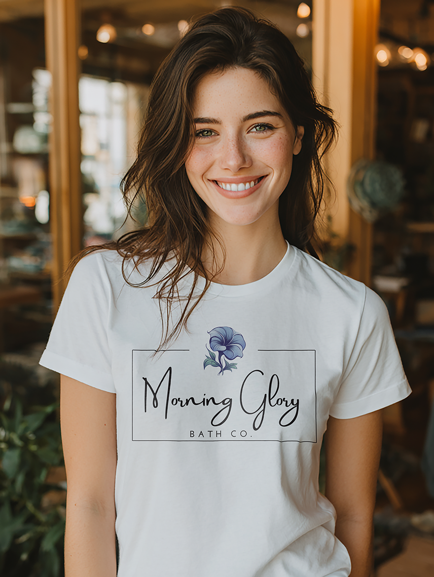 Smiling woman wearing a white Morning Glory Bath Co logo t-shirt in a sunlit indoor shop setting