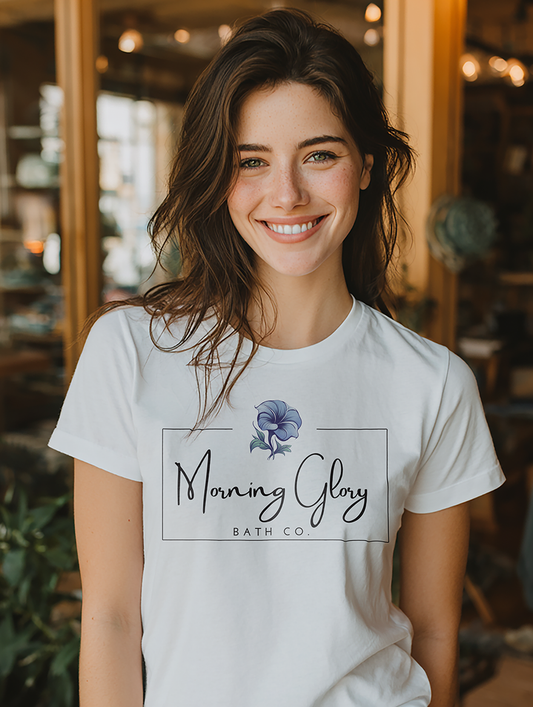 Smiling woman wearing a white Morning Glory Bath Co logo t-shirt in a sunlit indoor shop setting