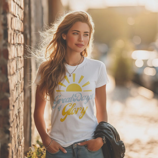 Woman wearing a white Morning Glory Bath Co t-shirt featuring a yellow sun graphic and the text 'Great Days of Glory' standing against a brick wall in warm sunlight video