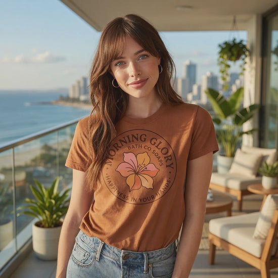 A woman wearing a caramel-colored Morning Glory Bath Co t-shirt with a circular floral logo and the words 'Happiness in Your Hands' on a coastal balcony overlooking the ocean video